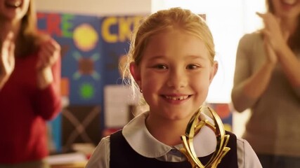 Happy graduating caucasian girl student holding golden trophy with blurred clapping women in school classroom
