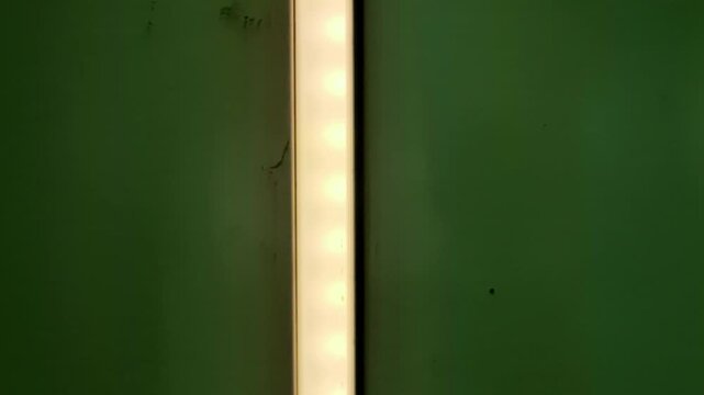 A vertical LED light bar with visible individual diodes glows with a warm, steady yellow light against a dark, forest-green wall that shows minor surface imperfections.