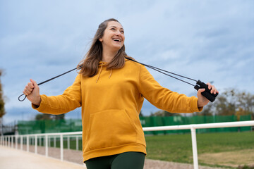 A woman in a yellow hoodie holds a jump rope and smiles against a sky background.