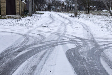 Tire tracks are visible on a snowy road in a rural setting during winter. The landscape shows signs of snowfall and trees