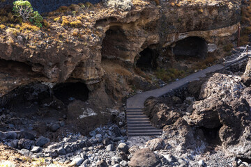 Volcanic sea caves carved into black basalt cliffs with stone staircase path amid dry grass at Costa El Sauzal, Tenerife, Canary Islands, Spain.