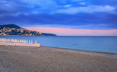 White umbrellas line a pebbled beach under golden sunset clouds in Nice, France