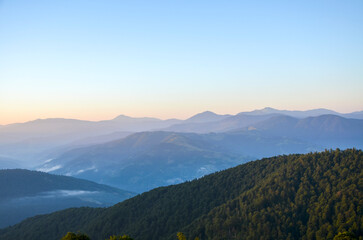 Tranquil dawn over layered mountain ridges, with forested slopes fading into a soft blue horizon. Mist drifts above valleys as the sun rises, creating peaceful scenery and a sense of vast openness
