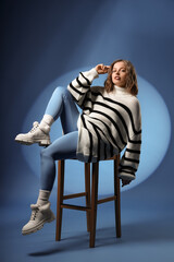 Stylish young woman with winter boots sitting in chair on blue background