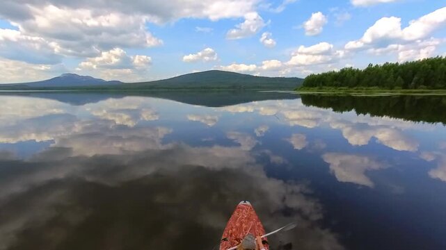A scenic wide view from the perspective of a small boat on a tranquil lake in the Southern Urals region of Russia. The image captures dramatic blue and white cumulus clouds perfectly mirrored in the s