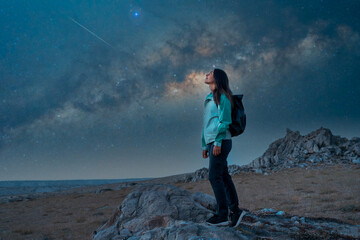 Woman standing on rocks, looking up at the starry night sky with a shooting star and Milky Way