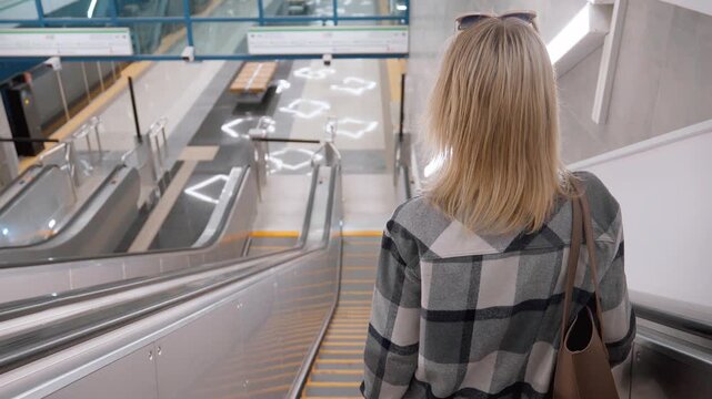Rear view of a young woman with blonde hair wearing a plaid shirt and carrying a shoulder bag, descending on an escalator inside a modern and empty subway station to the train platform