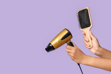 Female hands with modern hair dryer and brush on lilac background