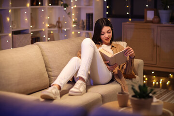 Obraz premium Beautiful young woman reading book and sitting on sofa in living room in evening