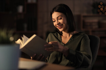 Happy young woman reading book at table in room at night