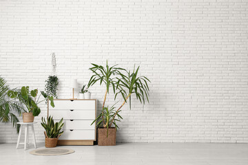 Interior of room with chest of drawers and houseplants near white brick wall