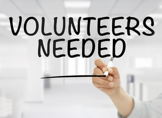 A conceptual business image showing "Volunteers needed" handwritten in bold black letters on transparent glass, with a blurred modern white office interior in the background. 