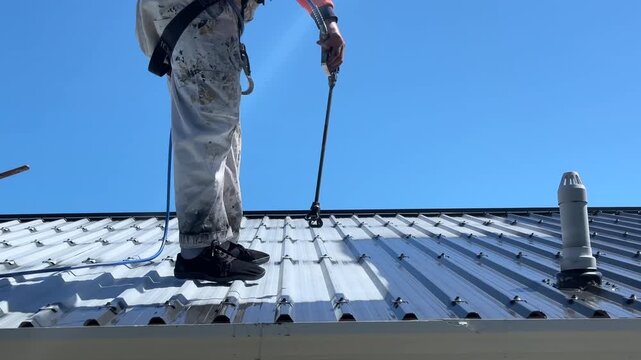 Workman in paint-splattered white overalls using a professional high pressure spray gun wand to repaint a long-run metal roof in a dark colour on a sunny day