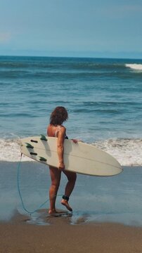 Portrait: Senior woman in black swimsuit holds white surfboard, summer vacation at ocean beach. Middle aged female has active lifestyle, sport lesson, surfing activity. Face of old lady in retirement
