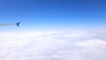 Aerial view from airplane window showing aircraft wing above white clouds and blue sky.