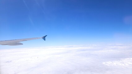 Fototapeta premium Aerial view from airplane window showing aircraft wing above white clouds and blue sky.