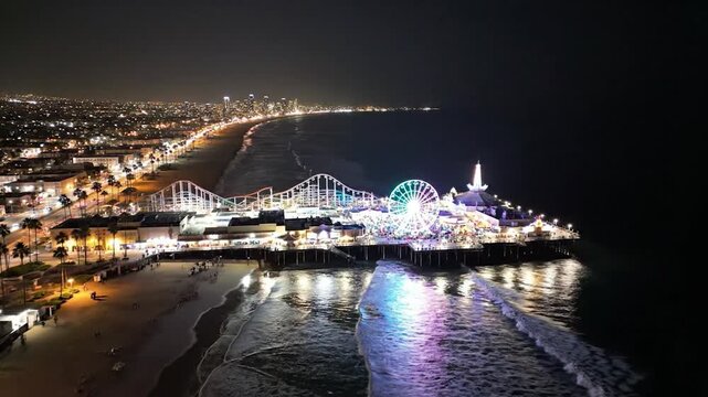 An aerial nighttime view of a brightly lit pier with a Ferris wheel, roller coaster, and beach