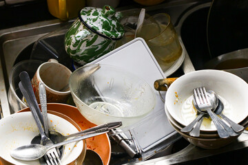 A pile of dirty dishes stacked in a kitchen sink with bowls, plates, spoons, and cups waiting to be...