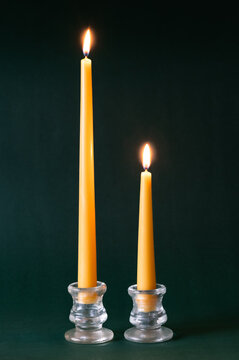 Two yellow beeswax taper candles burning in glass candle holders on dark green background