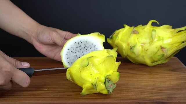 An Asian woman is cutting a yellow dragon fruit, scientific name Selenicereus megalanthus or Hylocereus megalanthus, into 2 parts with a knife, isolated on a black background.