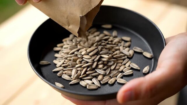 Close-up of hands pouring raw sunflower seeds from a paper bag into a black bowl, healthy snack