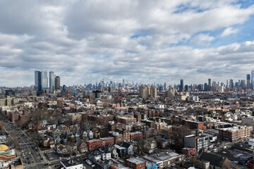Aerial drone photo from Jersey City featuring Manhattan skyline views, dense city blocks, waterfront buildings, and modern New York City architecture under dynamic daytime cloud cover.                