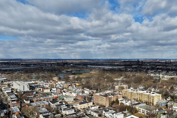 Aerial drone view over Lincoln Park in Jersey City with the Pulaski Skyway stretching across the horizon above the Hackensack River. Surrounding residential neighborhoods, open green space, and waterw