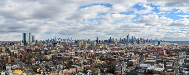 Panoramic aerial photo of Jersey City capturing a wide urban landscape with dense residential blocks, major streets, and the Manhattan skyline stretching across the horizon beneath dramatic, cloud-fil