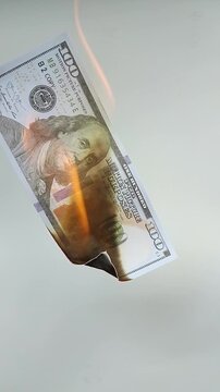 A close-up photo of a US one hundred dollar bill burning in midair against a clean, light background