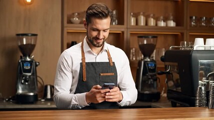 A smiling barista, wearing an apron, checks a smartphone while in a coffee shop