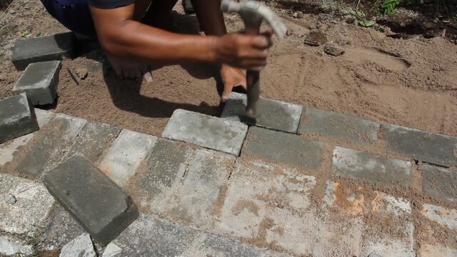 A construction worker interlocking paving stones on a sand bed. A hammer is used to level each paving stone.