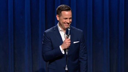 Man in suit holding a microphone, appearing to be on stage, with a blue curtain backdrop
