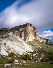 A majestic, white-faced mountain towers under a partly cloudy sky. Its rugged face contrasts with the verdant slopes below, revealing a winding path
