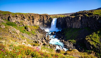 Obraz premium A majestic waterfall cascades down a rugged cliff, surrounded by lush green vegetation under a clear blue sky. The water flows through a rocky riverbed