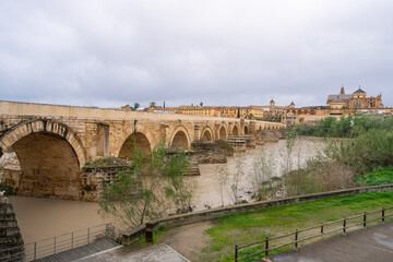 Ancient stone bridge with multiple arches crossing the Guadalquivir River in historic Cordoba, Andalusia, with the iconic Mosque-Cathedral visible.