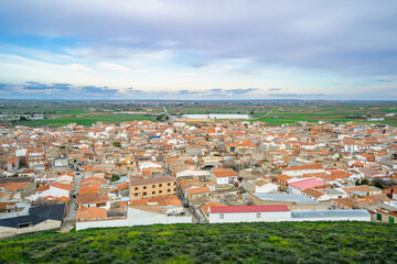 Elevated panoramic vista showcasing the traditional town of Consuegra surrounded by green agricultural plains under dramatic clouds along the Don Quixote Route.