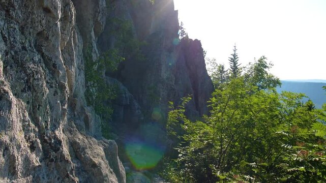 A sunlit view of a steep, rugged rock face and cliff, overlooking a dense green forest and a hint of water or sky in the distance. The shot captures the natural beauty and wilderness of the Southern U