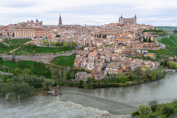 The ancient city of Toledo Spain glows softly at spring twilight with the Tagus River curving around the historic walls forming a natural barrier while lush greenery frames this elevated view.