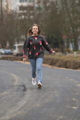 Fototapeta premium Young girl wearing a patterned sweater and jeans joyfully balances on a road, showcasing playful movement and carefree spirit in an urban environment with trees in the background