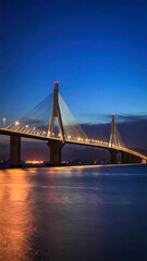 Fototapeta premium A nighttime long exposure shot of a majestic cable-stayed bridge, its lights reflected in the calm water below. The sky shows dusk