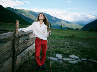Naklejka premium Woman hiking in a white sweater and red pants leans on a wooden fence in an expansive countryside with distant mountains, blue sky, and open landscape, embodying outdoor adventure and serenity.