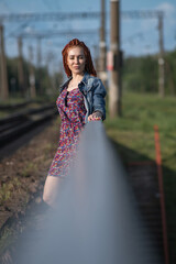 Young woman with long red hair wearing a floral dress and denim jacket walks along abandoned railway tracks surrounded by greenery, capturing a moment of carefree exploration