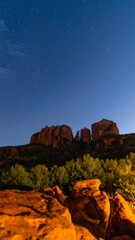 A nighttime panorama showcases a rock formation silhouette beneath a starry sky. Dark, blurred foreground leads to a forest line at the base