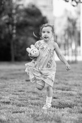 Young girl with a teddy bear running joyfully across a grassy field, wearing a light dress and sneakers, capturing the essence of childhood playfulness and freedom