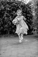Young girl with a teddy bear running joyfully across a grassy field, wearing a light dress and sneakers, capturing the essence of childhood playfulness and freedom