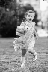 Young girl with a teddy bear running joyfully across a grassy field, wearing a light dress and sneakers, capturing the essence of childhood playfulness and freedom