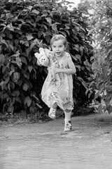 Young girl with a teddy bear running joyfully across a grassy field, wearing a light dress and sneakers, capturing the essence of childhood playfulness and freedom