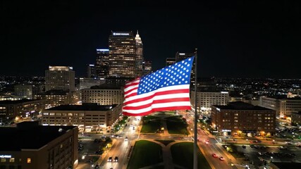 A patriotic American flag with red and white stripes and blue stars waves in the night wind against the iconic New York City skyline as a national symbol of freedom under the dark sky