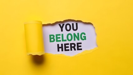 Torn yellow paper revealing the encouraging message 'YOU BELONG HERE' on a white background, symbolizing inclusion and welcome.