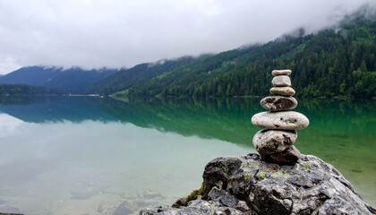 Balanced stone cairn on a rock by a serene mountain lake with misty forested hills and their reflections.